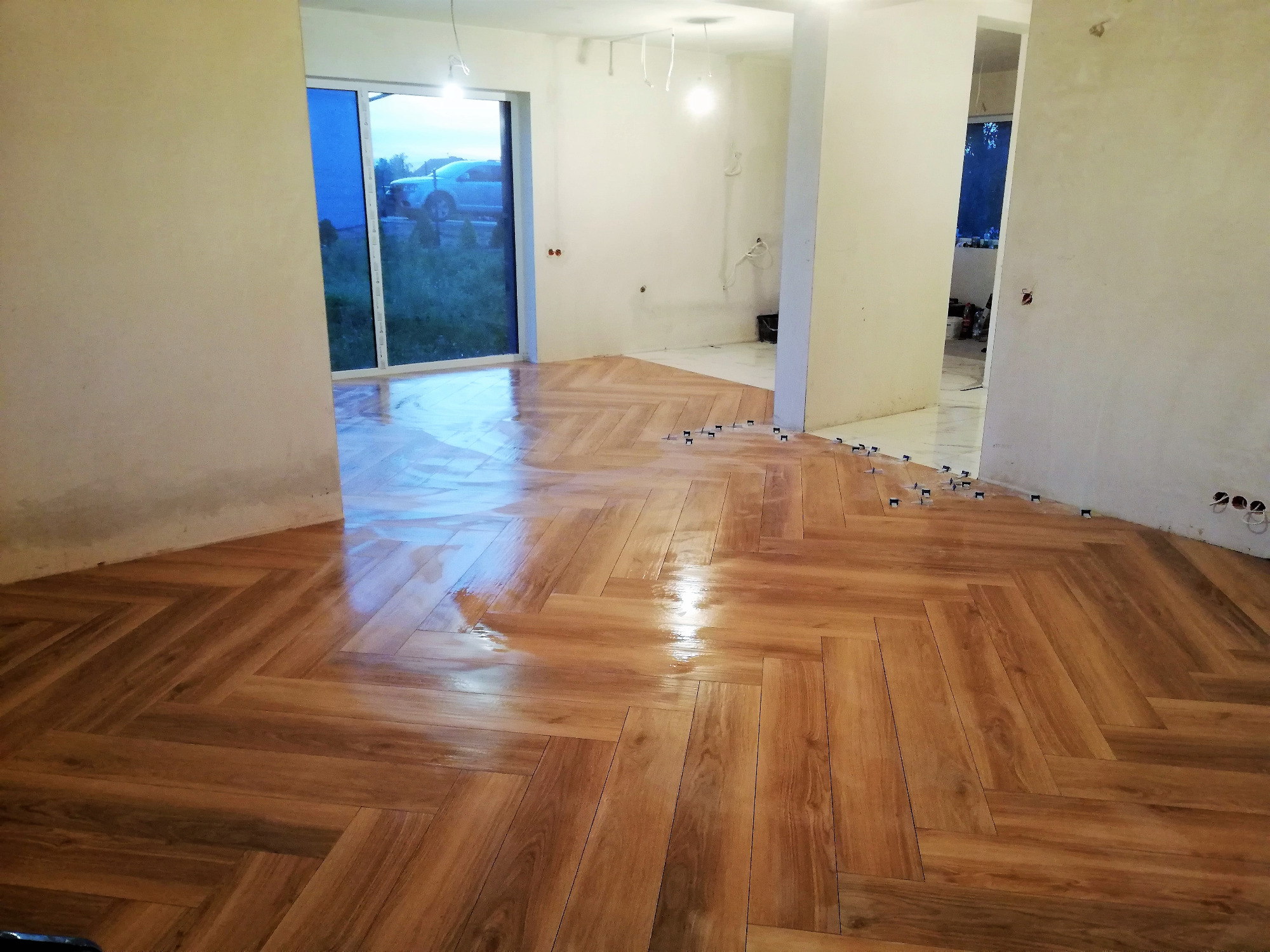 Układanie drewnopodobnych płytek w jodełkę na podłodze w trakcie remontu, widoczne krzyżyki dystansowe, ślady po kleju i okno z widokiem na samochód.