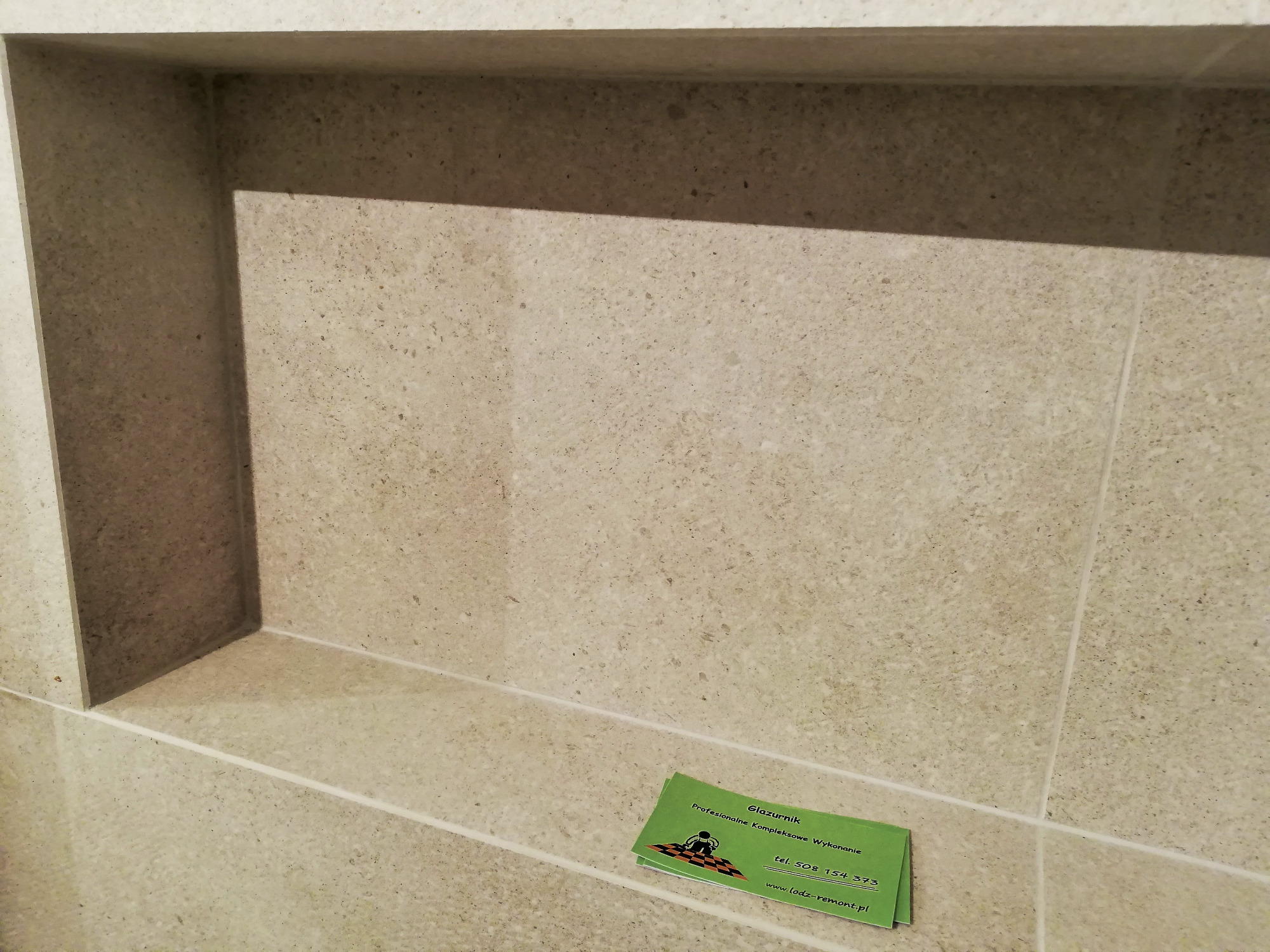 Wnęka w ścianie wyłożona beżowymi płytkami, na dolnej krawędzi leży wizytówka firmy glazurniczej.