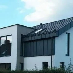 Nowoczesny dom z grafitowym dachem z blachy na rąbek stojący, białymi ścianami i dużymi oknami, widok z zewnątrz.