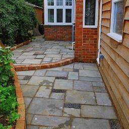 WOY-TECH Profesjonalne Budownictwo - Ułożona z szarych płyt chodnikowych ścieżka z wbudowanymi kratkami odpływowymi, obrzeżona czerwoną cegłą, przylegająca do drewnianej elewacji budynku.