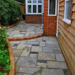 Ułożona z szarych płyt chodnikowych ścieżka z wbudowanymi kratkami odpływowymi, obrzeżona czerwoną cegłą, przylegająca do drewnianej elewacji budynku.