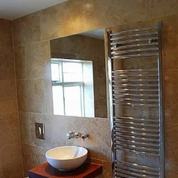 Wykończona łazienka z beżowymi płytkami imitującymi kamień, owalną umywalką nablatową na drewnianym blacie, chromowaną grzejnikiem drabinkowym i lustrem, w którym odbija się okno.