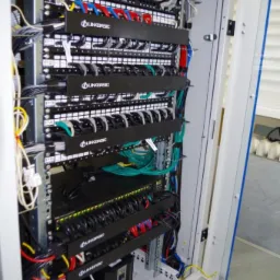 Szafa rack z panelami krosowniczymi Linkbasic i uporządkowanym okablowaniem LAN, widoczny zasilacz awaryjny UPS w dolnej części szafy.