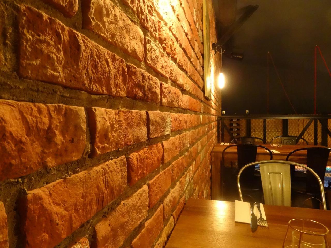 Ceglana ściana z wyraźną fakturą i oświetleniem punktowym w tle, widok na salę restauracyjną z drewnianymi stolikami i metalowymi krzesłami.