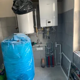 Montaż kotła gazowego Bosch i zasobnika Galmet, częściowo zakrytego niebieską folią ochronną, w pomieszczeniu wykończonym szarą glazurą. Widoczne rury instalacji CO i CWU w kolorze czerwonym...