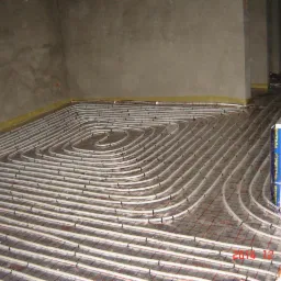 Układanie rur grzewczych na siatce w systemie ogrzewania podłogowego. Widoczne białe rury, szara siatka, surowy beton i fragment opakowania materiałów izolacyjnych.