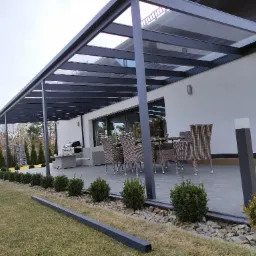 Nowoczesna metalowa pergola z przezroczystym zadaszeniem nad betonowym tarasem, z meblami ogrodowymi z rattanu, oświetleniem i starannie utrzymanym trawnikiem.