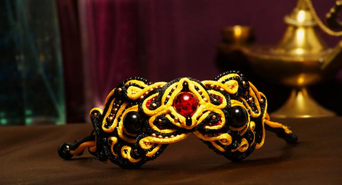 Czarno-złoty naszyjnik sutasz z czerwonym kamieniem centralnym i czarnymi koralikami, leżący na ciemnym blacie, z rozmytym tłem w odcieniach fioletu i złotą lampą Aladyna.