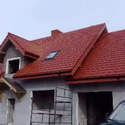 Dom w trakcie budowy z nowym, czerwonym dachem z dachówki metalowej i oknem dachowym, elewacja surowa, widoczne rusztowania.