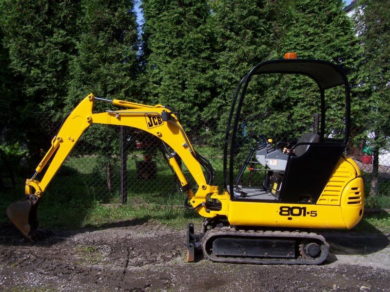 Żółta minikoparka JCB 801-5 na gąsienicach, zaparkowana na ziemi z zielonymi drzewami w tle.