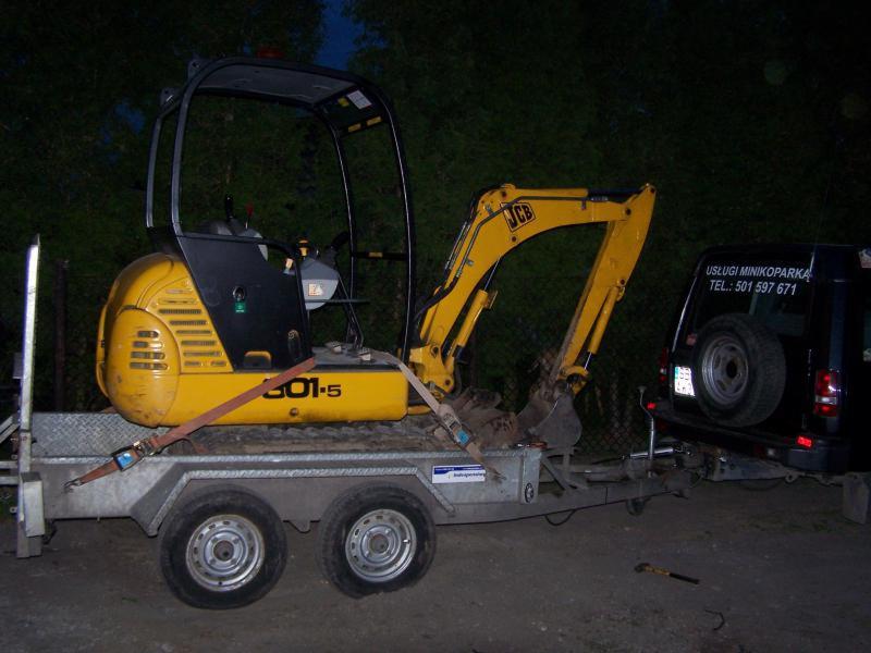 Żółta minikoparka JCB 801.5 zabezpieczona pasem transportowym na metalowej przyczepie, z tyłu zaparkowany czarny samochód z napisem 'Usługi Minikoparką' i numerem telefonu.