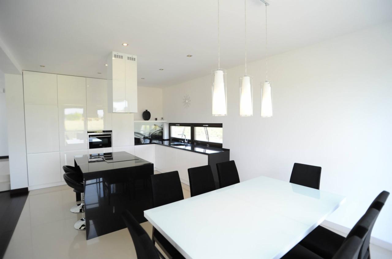 Nowoczesna, biała kuchnia z wysokim połyskiem, zabudowa szaf na wymiar z wyspą i czarnym blatem, uzupełniona białym stołem jadalnym i czarnymi krzesłami.