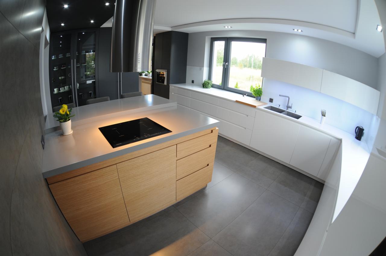 Nowoczesna kuchnia z drewnianą wyspą i płytą indukcyjną, białe szafki pod oknem, szare płytki podłogowe, oświetlenie punktowe, minimalistyczny design.