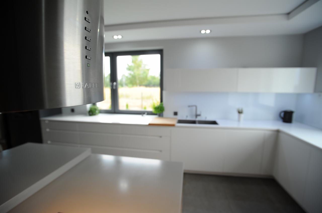 Minimalistyczna biała kuchnia z zabudową na wymiar, stalowy okap Faber, widok na okno z zielenią w tle.