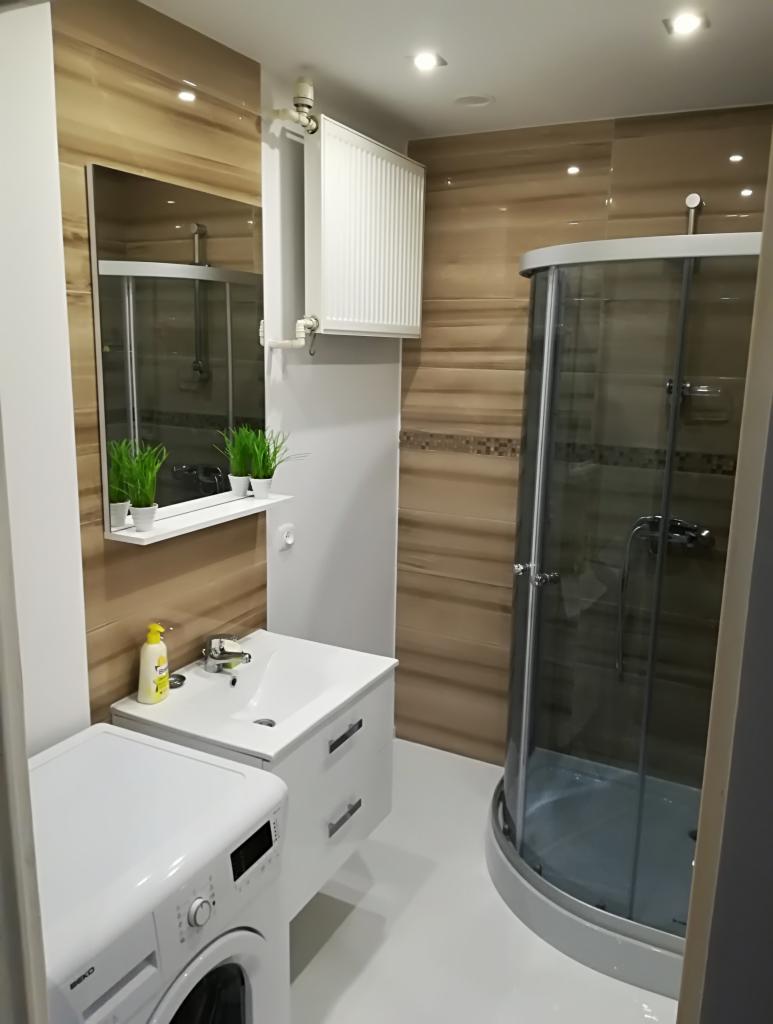 Ukończona łazienka z kabiną prysznicową, umywalką z szafką, pralką Beko i grzejnikiem na ścianie, wykończona brązowymi płytkami imitującymi drewno.
