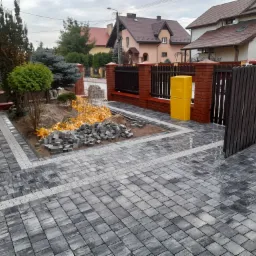 Ułożona szara kostka brukowa z jasnymi akcentami na podjeździe, obok przygotowany stos kostki, widoczne ogrodzenie z cegły i drewna, oraz elementy zieleni w ogrodzie.