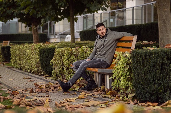 Mężczyzna w oliwkowej bluzie z kapturem i podartych dżinsach siedzi na ławce w parku, jesienne liście na chodniku, w tle budynek z balkonami.