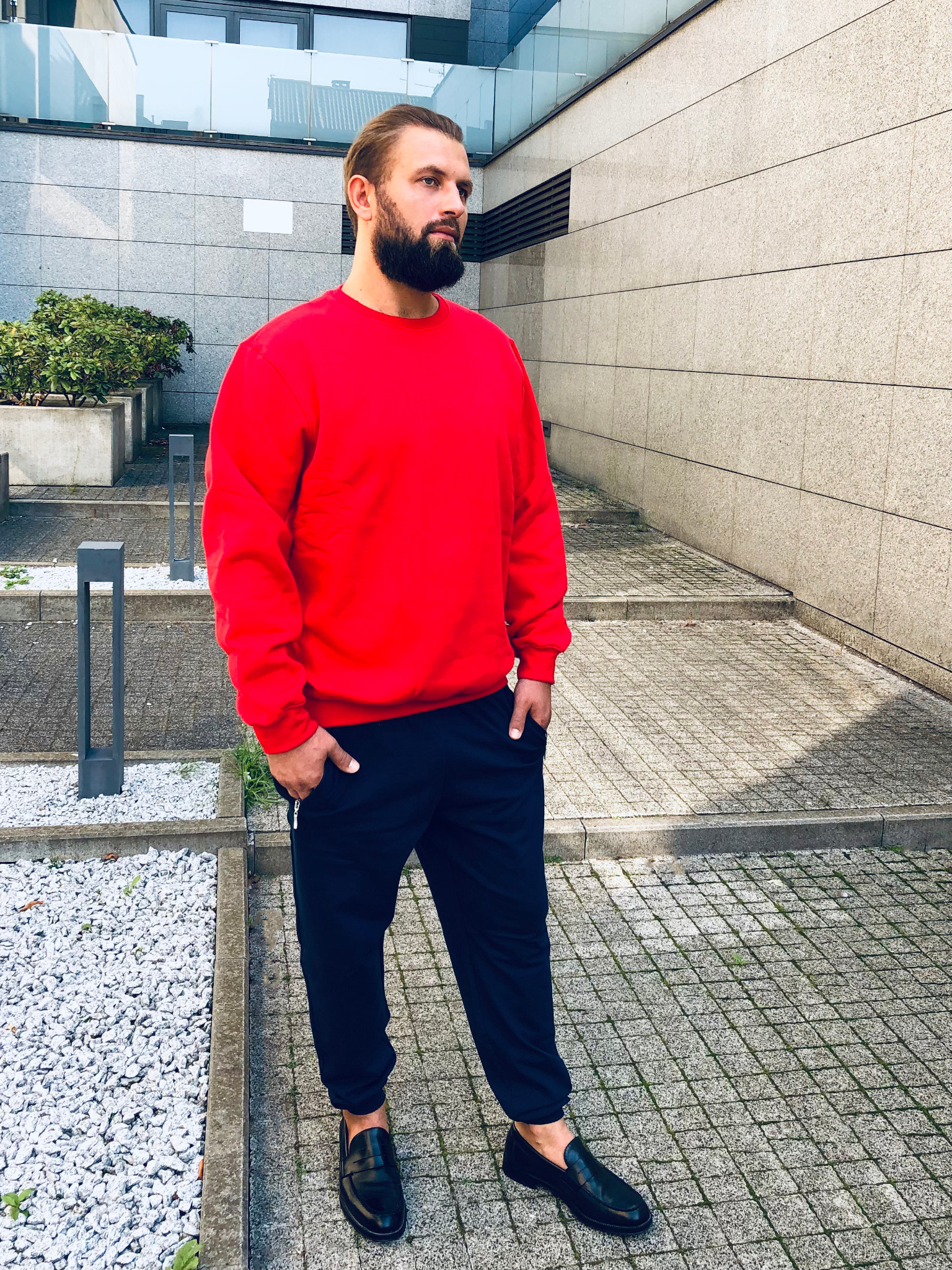 Mężczyzna z brodą w czerwonej bluzie i granatowych spodniach, pozuje na tle nowoczesnego budynku z kamienną elewacją.