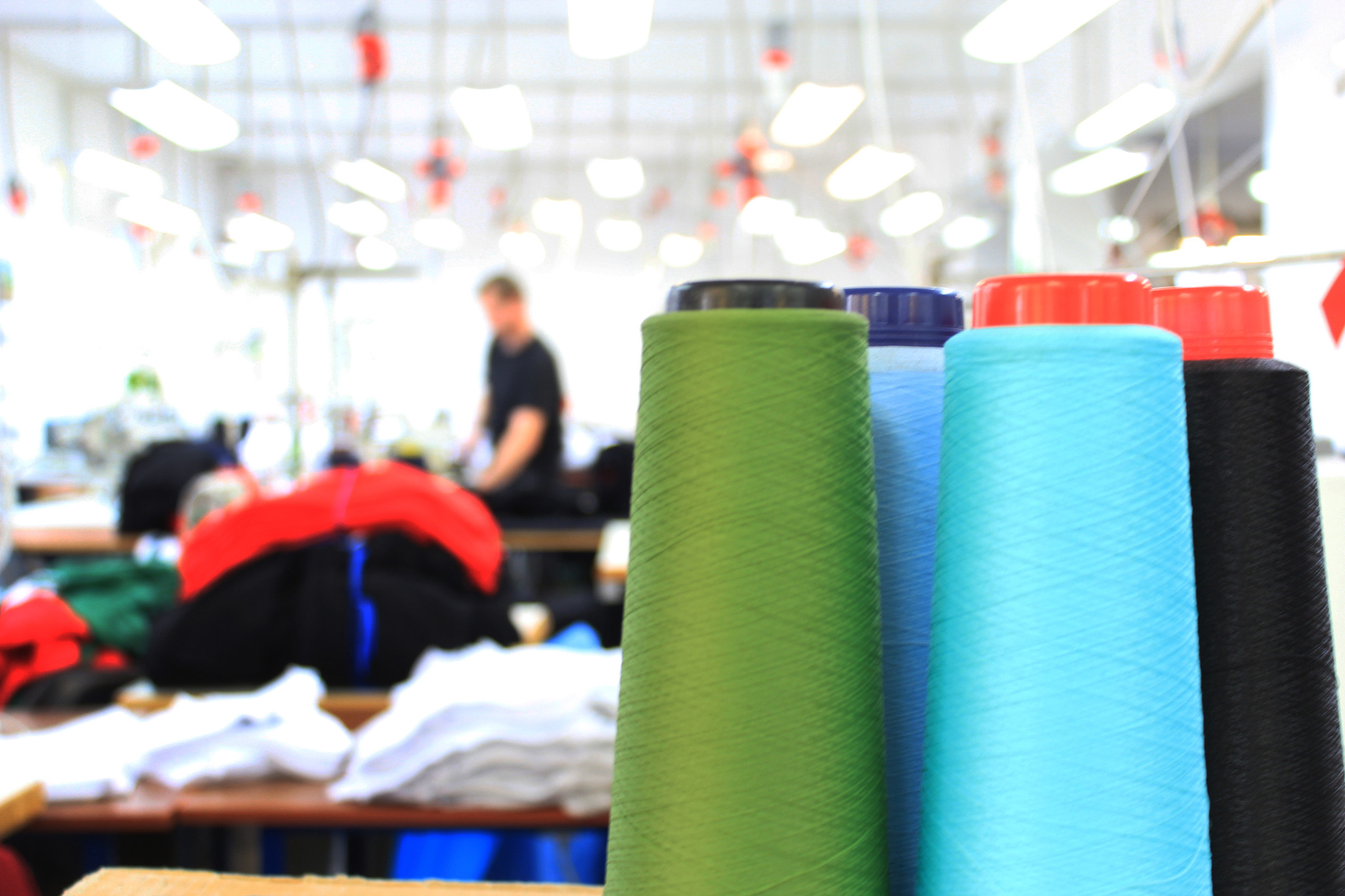 Kolorowe szpule nici w szwalni, w tle pracownik przy maszynie, stosy materiałów.