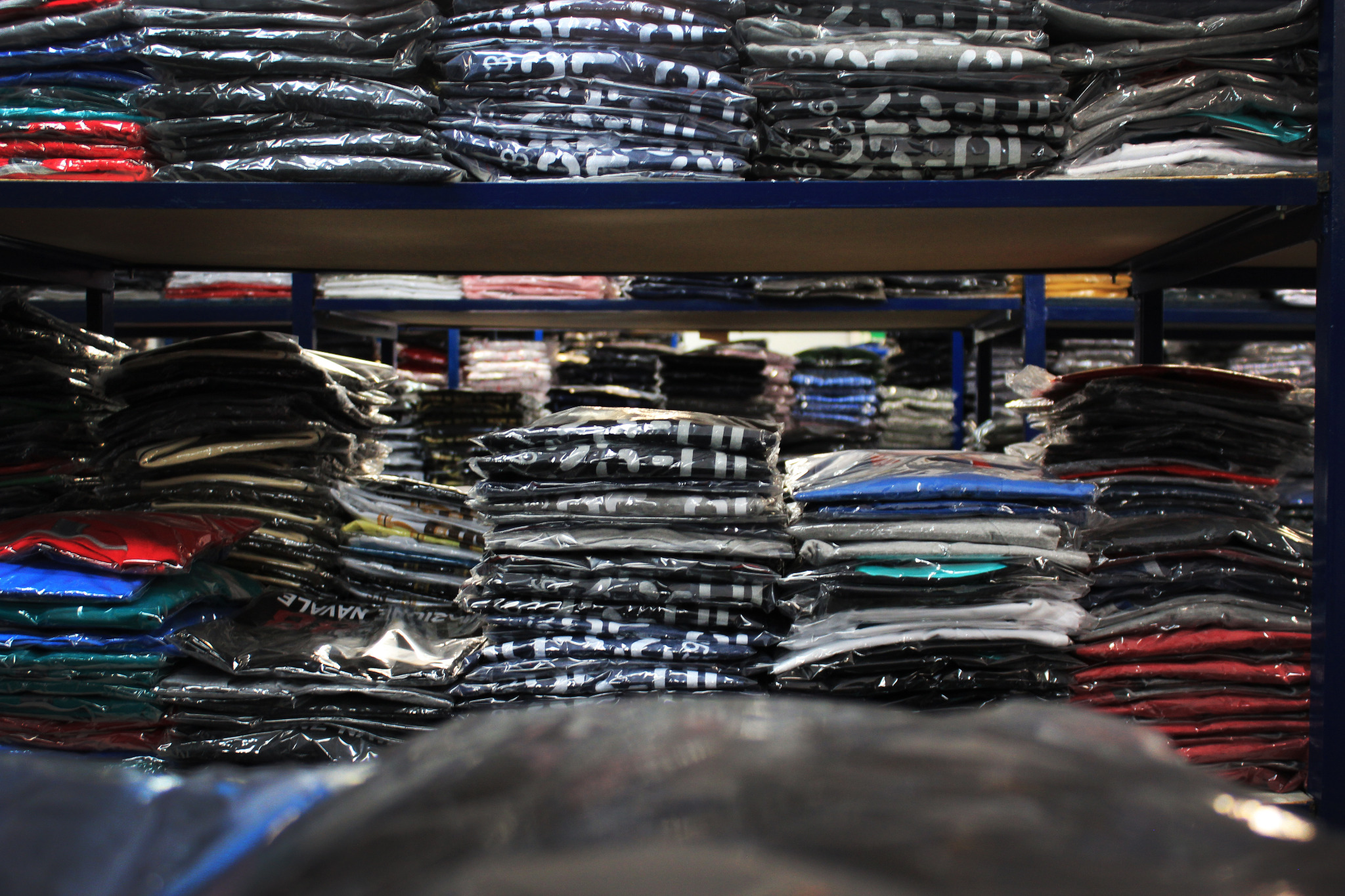 Stosy zapakowanych koszulek na metalowych regałach magazynowych, widoczne różne kolory i wzory, część w przezroczystej folii.