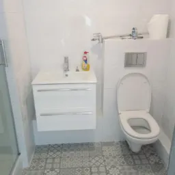 Wykończona łazienka z białą armaturą: wisząca szafka z umywalką, toaleta z podtynkowym systemem spłukiwania, białe płytki na ścianach i wzorzysta szaro-biała podłoga.