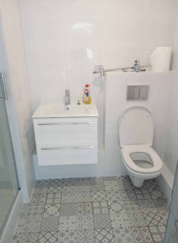 Wykończona łazienka z białą armaturą: wisząca szafka z umywalką, toaleta z podtynkowym systemem spłukiwania, białe płytki na ścianach i wzorzysta szaro-biała podłoga.