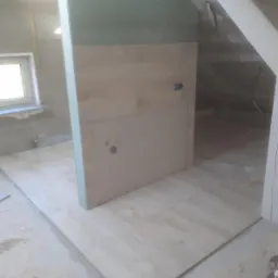 Wnętrze poddasza w trakcie remontu: częściowo wykończona podłoga z jasnych paneli, fragment ściany z płyt kartonowo-gipsowych i widoczne elementy konstrukcji dachu.