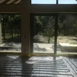 Rozłożona instalacja ogrzewania podłogowego na folii izolacyjnej w surowym wnętrzu domu z dużymi oknami i widokiem na zieleń.