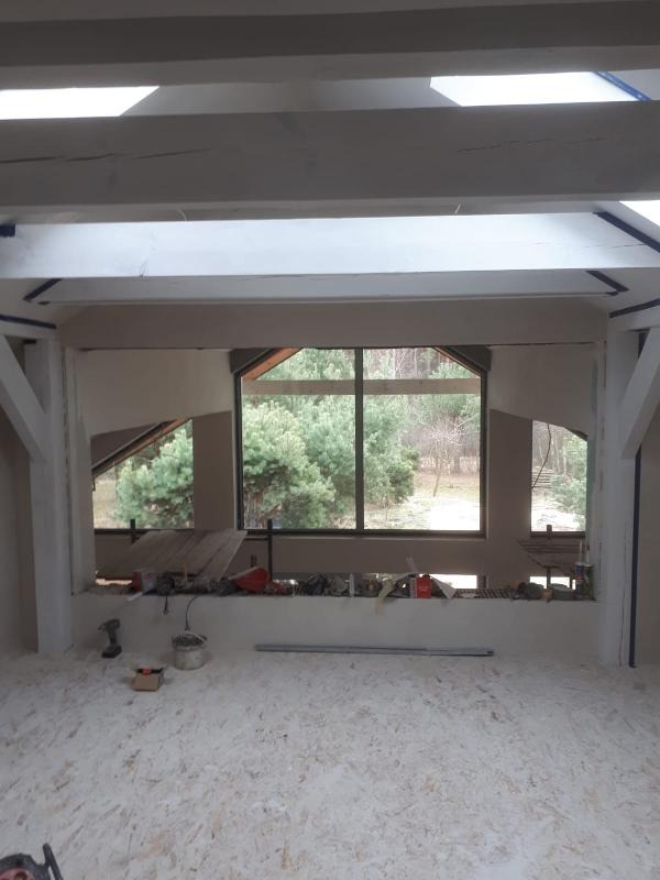 Wnętrze w trakcie remontu: widok na okno z drewnianą ramą, surowy parkiet na podłodze, widoczne narzędzia i materiały budowlane, białe belki stropowe.