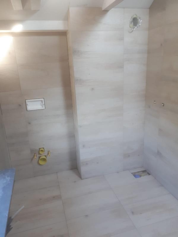 Wykończona łazienka z beżowymi płytkami imitującymi drewno, widoczne przyłącza wodno-kanalizacyjne i otwór w podłodze.