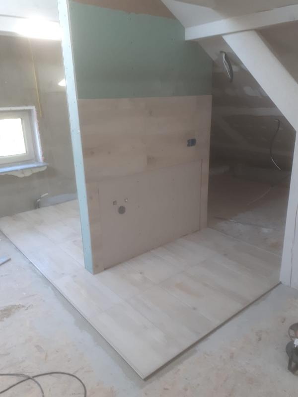 Wnętrze poddasza w trakcie remontu: częściowo wykończona podłoga z jasnych paneli, fragment ściany z płyt kartonowo-gipsowych i widoczne elementy konstrukcji dachu.