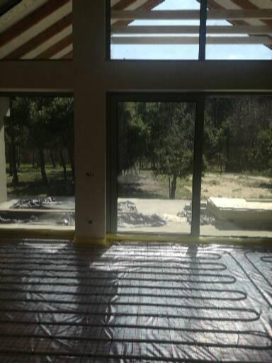 Rozłożona instalacja ogrzewania podłogowego na folii izolacyjnej w surowym wnętrzu domu z dużymi oknami i widokiem na zieleń.