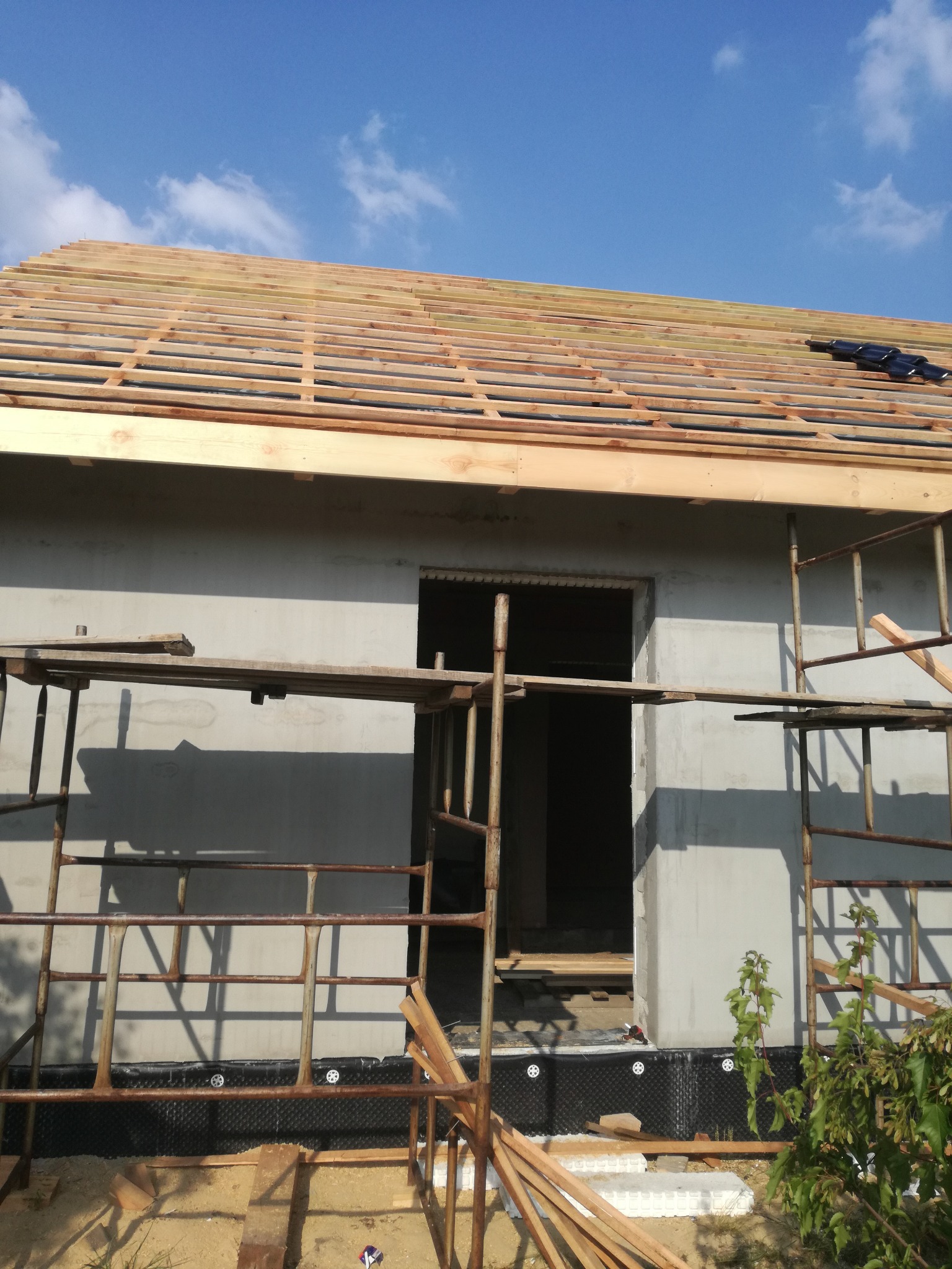 Surowy stan budowy domu jednorodzinnego: widok na szkielet dachu z drewnianych łat i kontrłat, fragment ściany z otworami okiennymi, rusztowania i czarna folia fundamentowa z białymi kołkami...