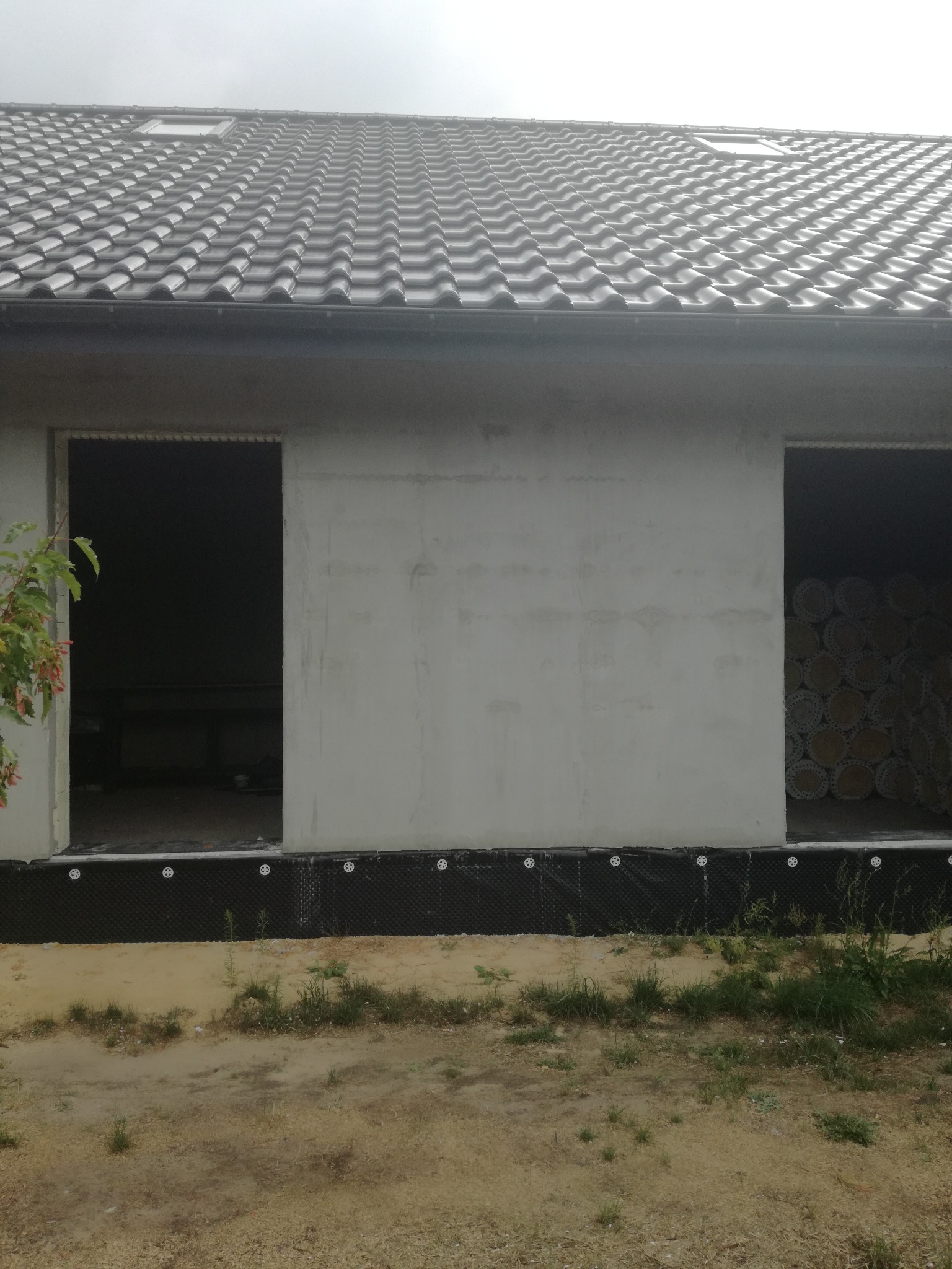 Surowy stan budynku w Tarnowskich Górach z widoczną izolacją fundamentów i przygotowanymi otworami okiennymi, dach pokryty ciemną dachówką. W jednym z otworów widoczne rolki materiału izolacyjnego.