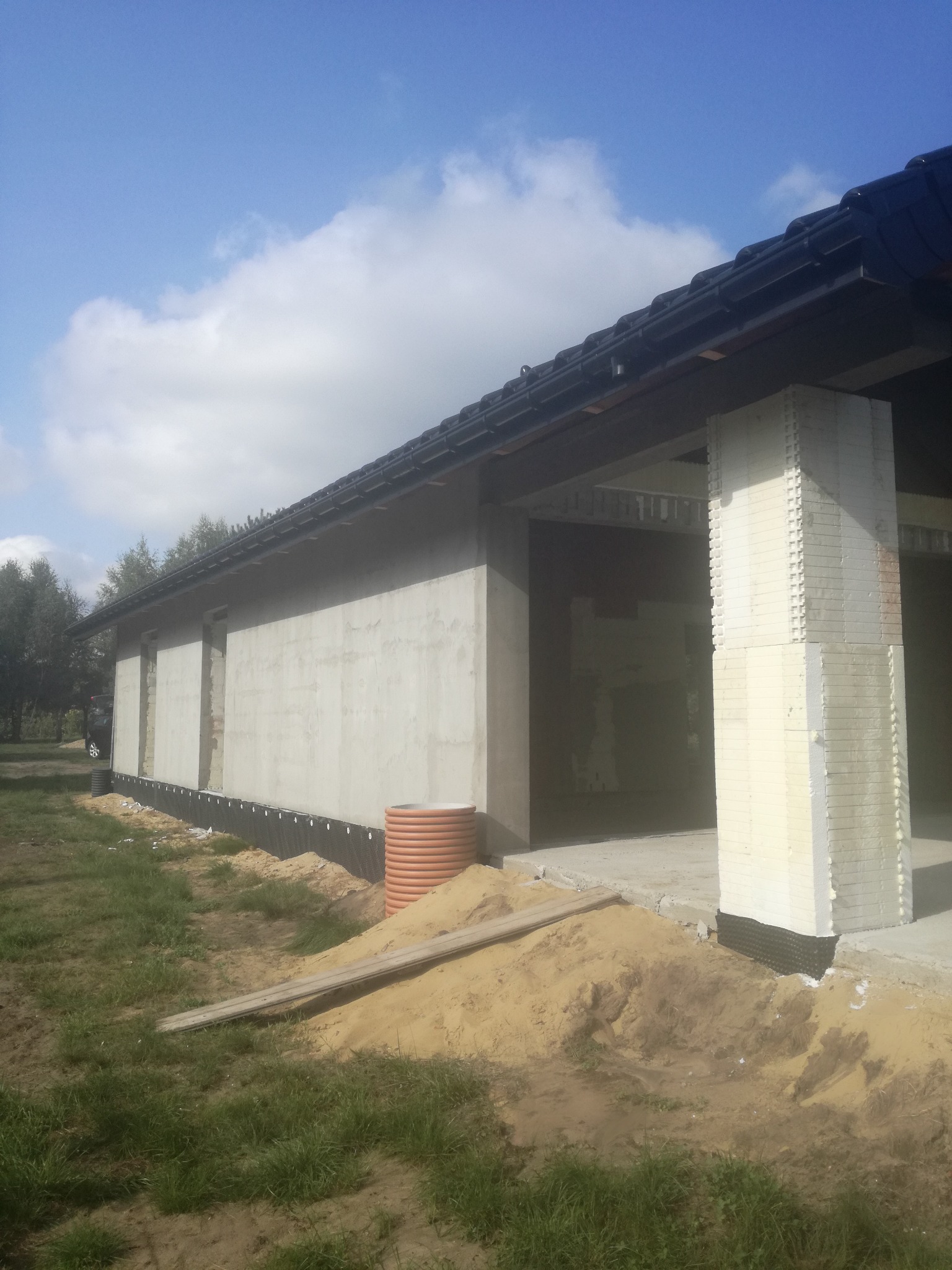 Surowy stan budynku jednorodzinnego w trakcie budowy, widoczne fundamenty z izolacją, ściany z betonu komórkowego, kolumna z izolacji termicznej, przewód kanalizacyjny w wykopie, oraz niebo...