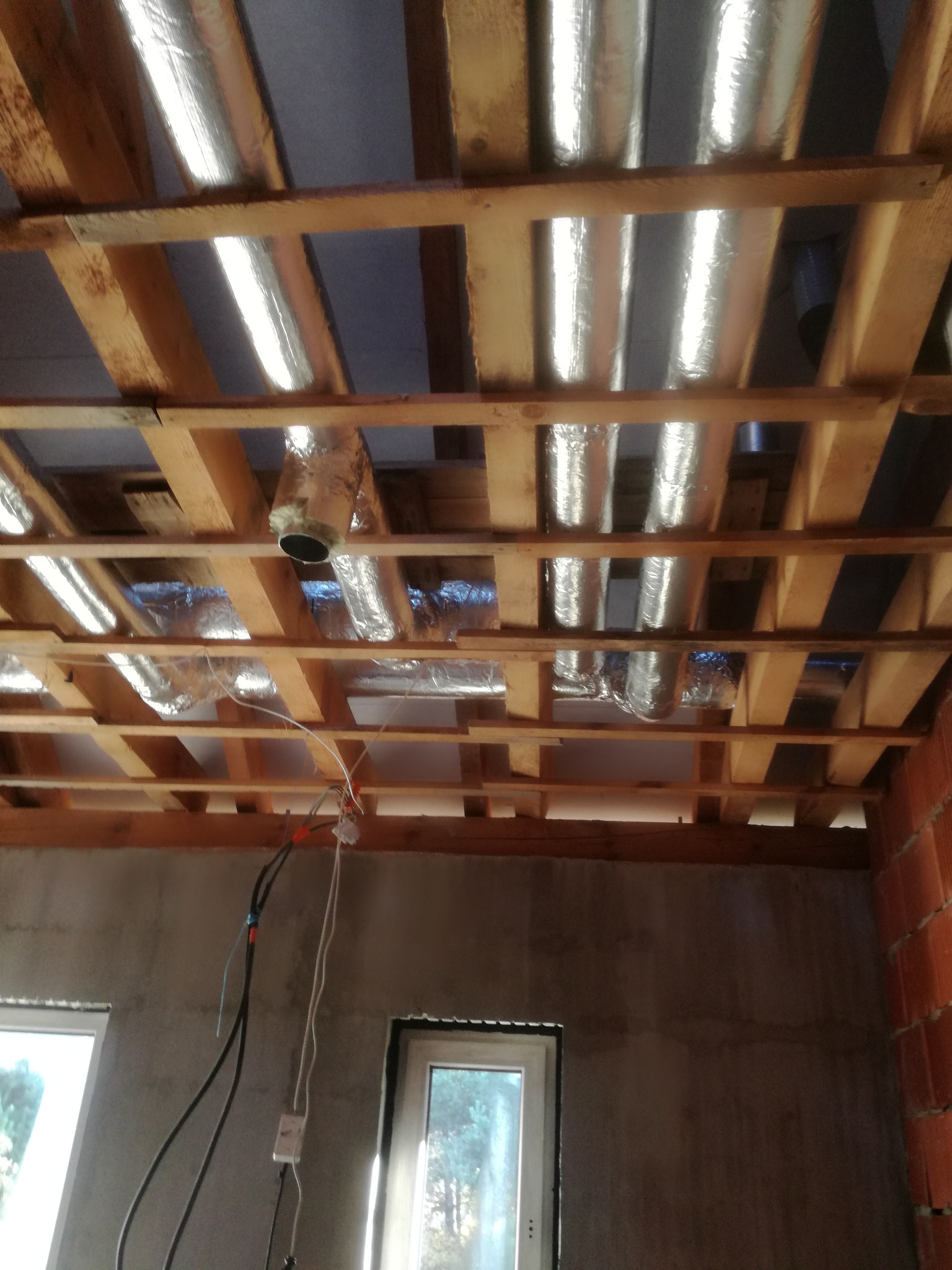 Widok od dołu na drewniany szkielet sufitu z izolowanymi, aluminiową folią przewodami wentylacyjnymi, widoczne okna i kable elektryczne zwisające ze ściany.