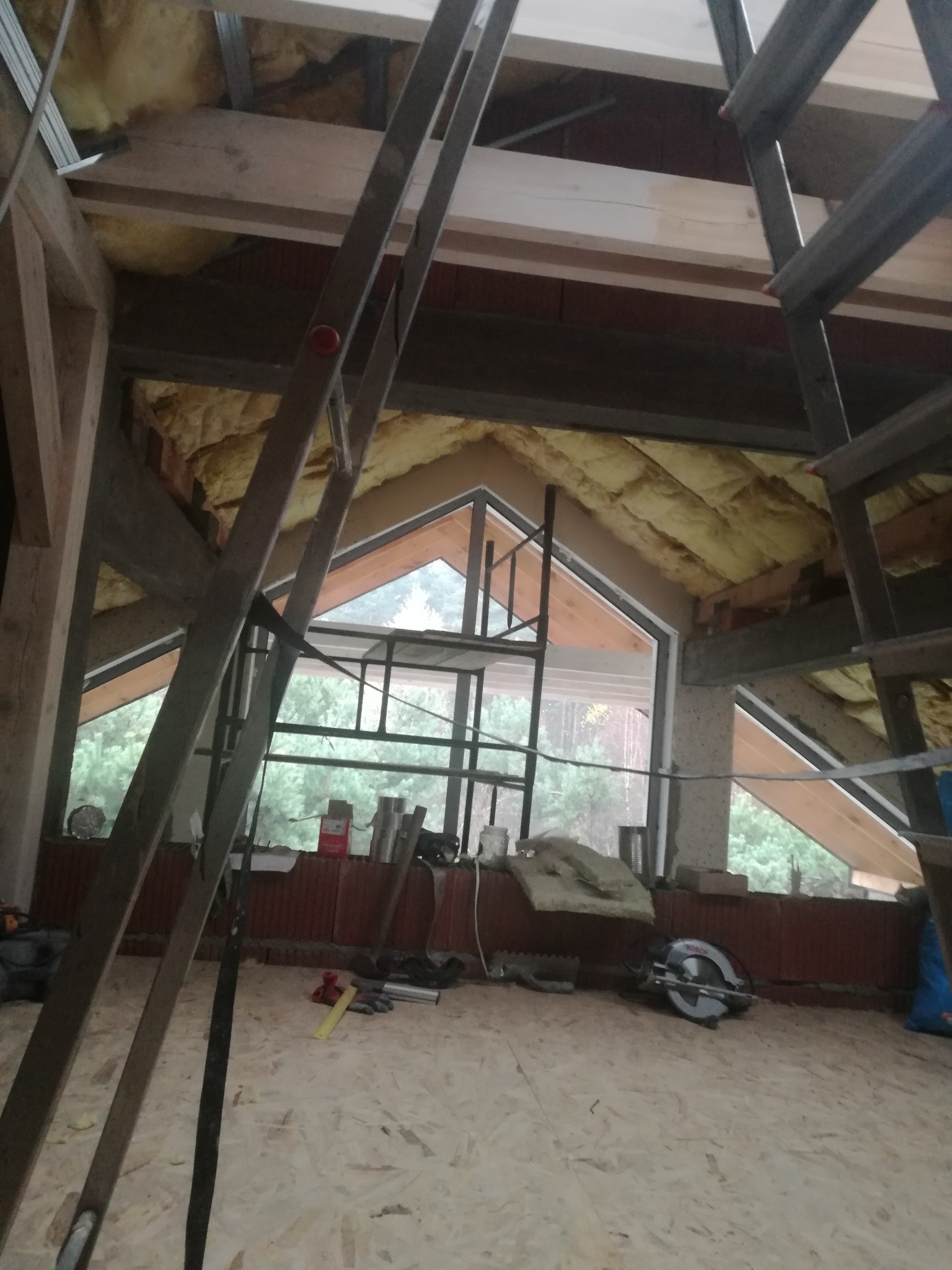 Montaż okna dachowego w nowym domu, widoczne ocieplenie wełną mineralną, konstrukcja drewniana, drabina, narzędzia budowlane i piła tarczowa na podłodze z płyt OSB.