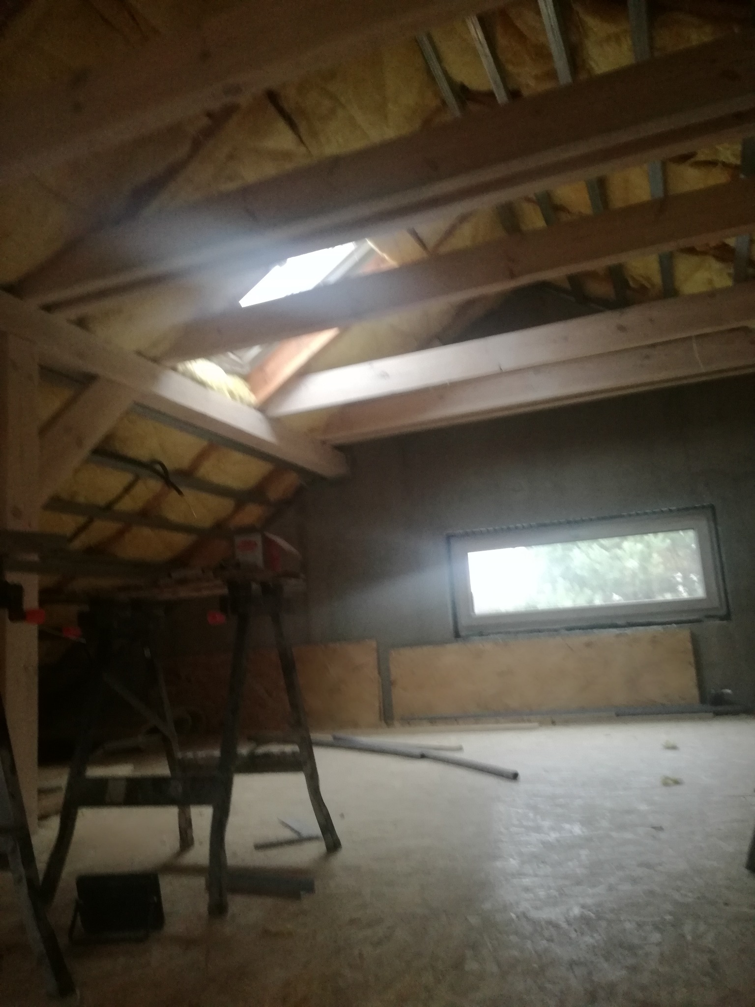 Wnętrze poddasza w trakcie adaptacji: widoczne drewniane krokwie, izolacja z wełny mineralnej, okno dachowe i fragment ściany z oknem. Na podłodze widoczne rury i resztki materiałów budowlanych.