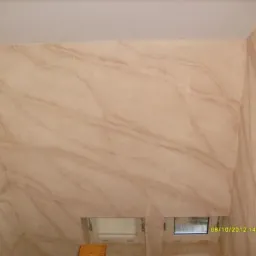 Ściana w łazience wykończona dekoracyjnym tynkiem imitującym marmur w odcieniach beżu i brązu, widoczne dwa małe okna i fragment drewnianego krzesła.