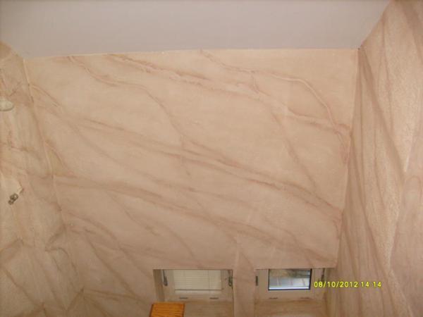 Ściana w łazience wykończona dekoracyjnym tynkiem imitującym marmur w odcieniach beżu i brązu, widoczne dwa małe okna i fragment drewnianego krzesła.