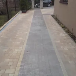 Ułożona kostka brukowa w odcieniach beżu i szarości tworzy ścieżkę wzdłuż domu, prowadzącą do bramy. Widoczne elementy ogrodu i zaparkowany samochód w tle.