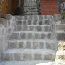 Kamienne schody zewnętrzne, prowadzące w górę, wykonane z jasnoszarego kamienia o nieregularnej fakturze, z widocznymi spoinami i fragmentem drewnianej konstrukcji w tle.