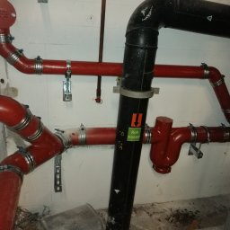 Instal-Mat - Czerwone i czarne rury instalacji wodnej z metalowymi obejmami zamocowane do betonowej ściany, widoczny fragment instalacji z zaworem i napisem 'Roh wasser'.