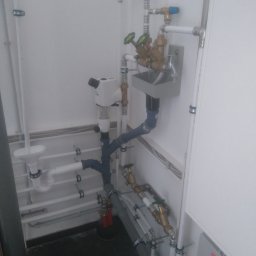 Instal-Mat - Kompleksowa instalacja hydrauliczna z białymi rurami PCV, zaworami z zielonymi pokrętłami, filtrem wody z przezroczystym zbiornikiem i odpływem lejkowym, zamontowana w pomieszczeniu z białą ścianą...