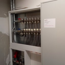 Instal-Mat - Rozdzielacz ogrzewania podłogowego w szafce, widoczne rury instalacyjne i zawory, na ścianie przyklejona ulotka informacyjna.