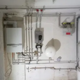 Biały piec gazowy Vaillant zamontowany na szorstkiej ścianie piwnicy z widocznymi rurami miedzianymi i instalacją, w tle licznik gazu i inne elementy instalacji.
