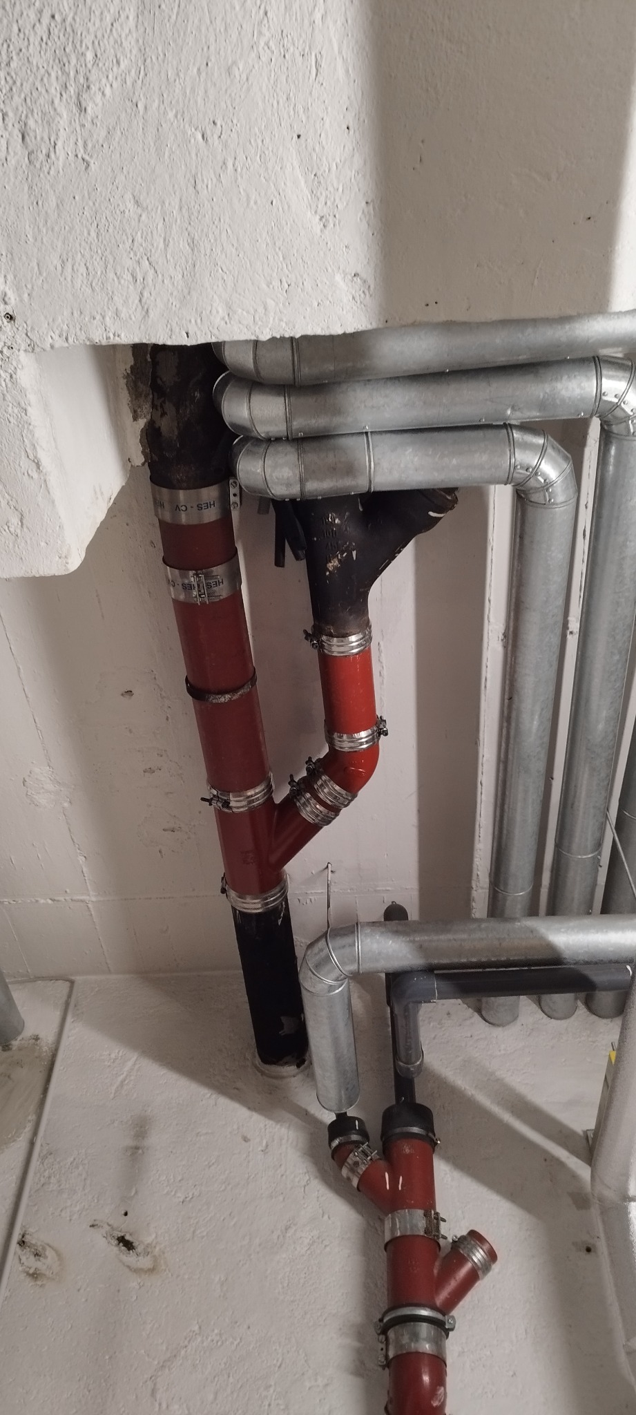 Złożony system rur hydraulicznych z czerwonymi i srebrnymi elementami, zamontowany przy białej, nierównej ścianie.