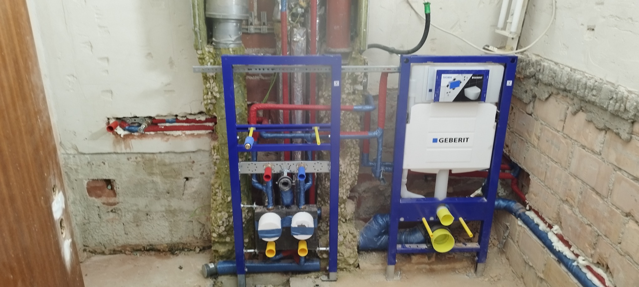 Stelaż podtynkowy Geberit z widocznymi rurami instalacji wodnej i kanalizacyjnej w surowym stanie, przed zabudową, z fragmentami odsłoniętej cegły i pianką montażową w tle.