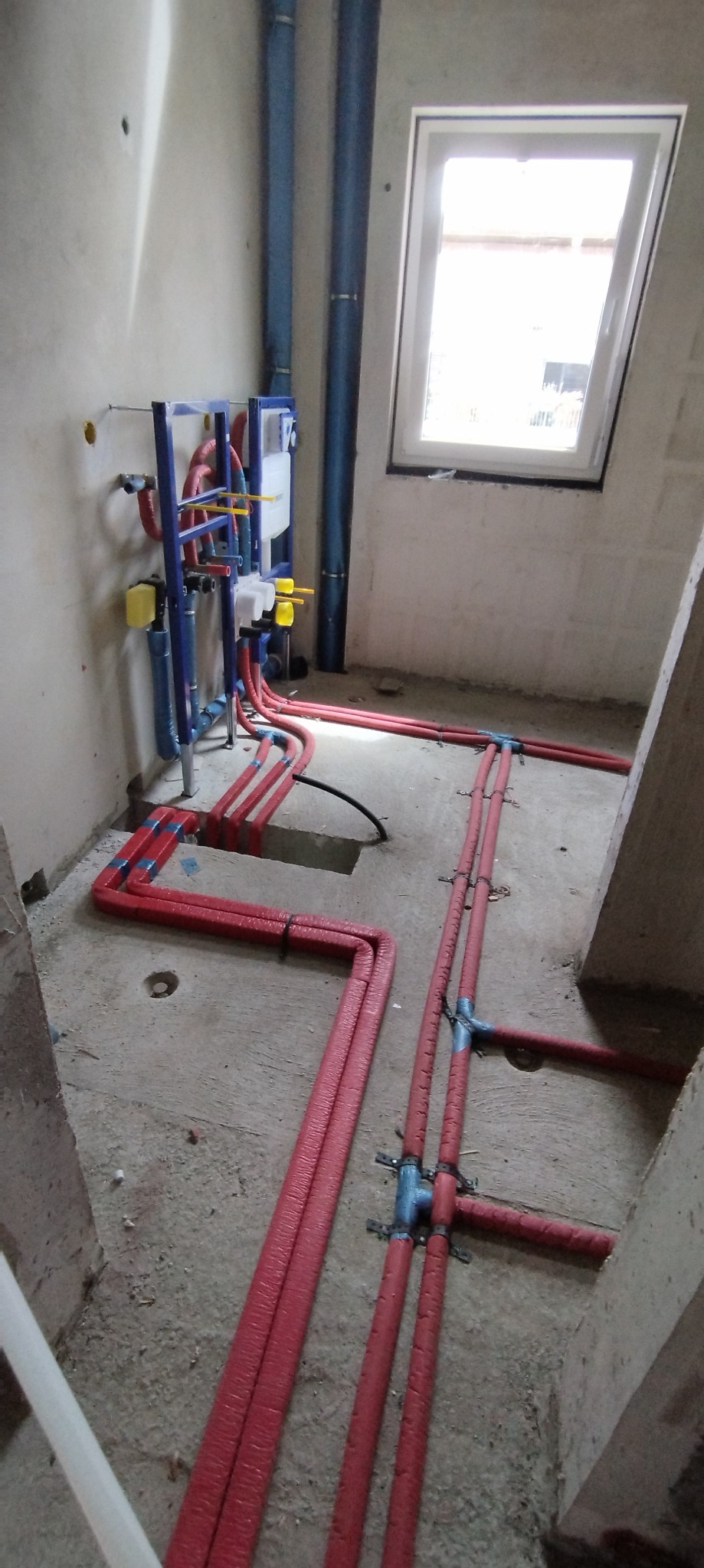 Wnętrze łazienki w trakcie remontu z widoczną instalacją hydrauliczną: czerwone rury z izolacją termiczną prowadzone po podłodze i niebieskie rury pionowe przy ścianie z zamontowaną podtynkową...