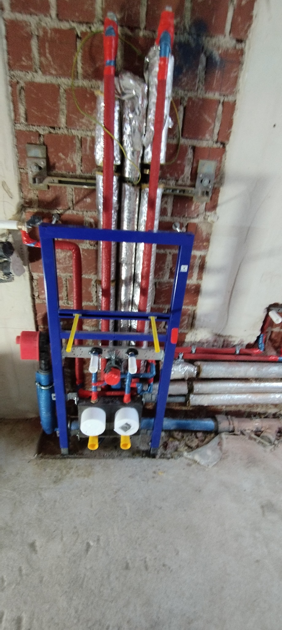 Stelaż podtynkowy do WC zamontowany przy ceglanej ścianie, widoczne rury z izolacją termiczną, instalacja hydrauliczna w trakcie montażu.